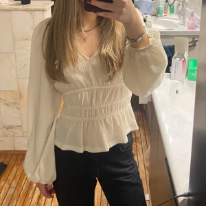 Vit blus med volangdetaljer - Säljer en stilren vit blus med v-ringning och volangdetaljer. Blusen har långa ärmar och en smickrande passform med resår i midjan. Perfekt för en chic look!