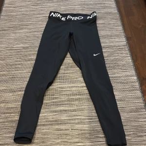 Svarta tights från Nike - Snygga svarta Nike Pro tights med hög midja och elastisk linning. Perfekta för träning med en tight passform och Nike-logga på benet. Materialet är stretchigt och bekvämt för maximal rörelsefrihet.