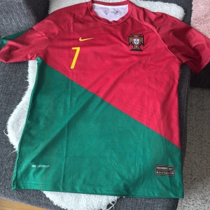 Röd/grön Portugal fotbollströja - Säljer en röd/grön Portugal fotbollströja från Nike med nummer 7 och Ronldos namn på baksidan. Tröjan har korta ärmar och ett broderat emblem på bröstet. Perfekt för fotbollsfans! Storlek L
