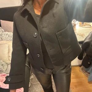 Svart croppad kavaj - Snygg svart croppad kavaj med stora knappar framtill. Jackan har långa ärmar och en klassisk krage. Perfekt för en stilren look. Andvänt 1 gång.