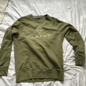 Olivgrön sweatshirt från Gant - Säljer en snygg olivgrön sweatshirt från Gant med deras ikoniska logga på bröstet. Tröjan har långa ärmar och ribbade muddar vid ärmslut och nederkant. Perfekt för en avslappnad stil.