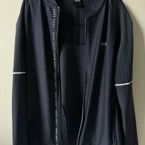  Hugo Boss - Snygg marinblå jacka från Hugo Boss med diskreta ränder på ärmarna och dragkedja framtill. Jackan har en stilren design med vita detaljer vid ärmsluten och logotyp på bröstet. Perfekt för en stilren look.