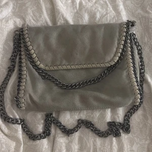 Grå axelväska med kedjedetaljer - Snygg grå axelväska i mocka med coola kedjedetaljer runt kanterna. Väskan har ett leopardmönstrat foder och en praktisk dragkedja. Perfekt för att ge din outfit en edgy touch en liten fläck på baksidan har försökt få bort men går ej. Väskan liknar Stella McCartney väskan köpdes för 600. Priset kan diskuteras vid snabb affär