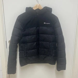 Svart pufferjacka från Champion - Säljer en svart pufferjacka från Champion. Jackan har en dragkedja framtill och en huva för extra skydd mot väder. Tillverkad i polyester med en mjuk och varm fyllning. Perfekt för kyligare dagar.