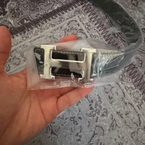 Svart bälte med silverfärgat spänne - Snyggt svart bälte med ett stilrent silverfärgat H-format spänne. Perfekt för att ge en elegant touch till din outfit. Bältet är inslaget i plast vilket ger en känsla av exklusivitet.