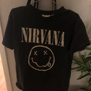 Svart Nirvana t-shirt - Cool svart t-shirt med Nirvana-tryck och ikoniskt smiley-motiv på framsidan. Perfekt för fans av klassisk rock. T-shirten har en normal passform och korta ärmar.