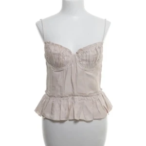 Beige korsettopp från Brock Collection x H&M - Säljer en elegant beige korsettopp från Brock Collection x H&M. Toppen har smala axelband och en volangdetalj vid nederkanten. Den har en dragkedja i ryggen för enkel påklädning. Perfekt för en stilren look.