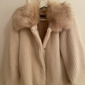 Beige/krämvit kort kappa med pälsdetaljer - Säljer en elegant beige kappa med en lyxig pälskrage. Jackan är tillverkad i Italien och har en stilren design med långa ärmar och en mjuk, ribbad textur. Perfekt för att hålla sig varm och snygg under kyliga dagar. 🧥✨Aldrig använd och köpt i en fin butik i Barcelona i höstas! Köpt för 1000kr men säljer för 790kr💫 