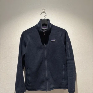 Mörkblå fleecejacka från Patagonia - Säljer en mörkblå fleecejacka från Patagonia med dragkedja framtill och en liten logga på bröstet. Jackan har långa ärmar och en praktisk ficka med dragkedja på ärmen. Perfekt för kyliga dagar!