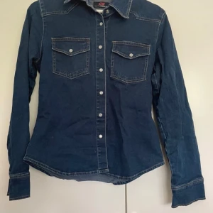 Mörkblå jeansskjorta med knappar - Snygg mörkblå jeansskjorta från C&A med långa ärmar och stilrena knappar framtill. Skjortan har två bröstfickor med lock och knappdetaljer. 