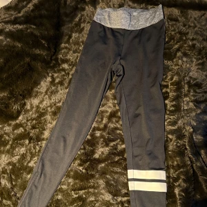 Svarta leggings med text och ränder - Snygga svarta leggings med grå midja och vita ränder vid vristen. Texten 'DEDICATION' på ena benet. Perfekta för träning eller en avslappnad dag. De har en dragkedja vid vristen