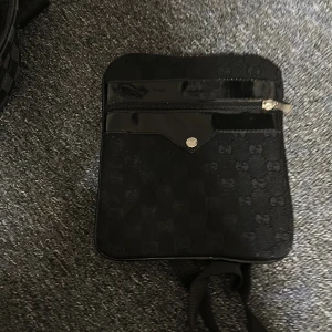 Axelväska  - Snygg svart axelväska med diskret mönster och dragkedja framtill. Väskan har en stilren design med en knappdetalj och är perfekt för att bära dina nödvändigheter.