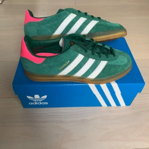 Adidas Gazelle i grönt och rosa - Snygga Adidas Gazelle sneakers i grönt mocka med vita ränder och en cool rosa detalj på hälen. Skorna har en klassisk design med snörning och en gummisula i brunt. Perfekta för en stilren look.