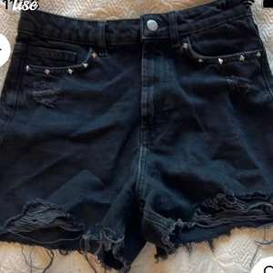 Svarta jeansshorts med nitar - Snygga svarta jeansshorts med slitna detaljer och coola nitar vid fickorna. De har en högmidjad passform och fransiga kanter som ger en avslappnad look. Perfekta för en trendig stil!