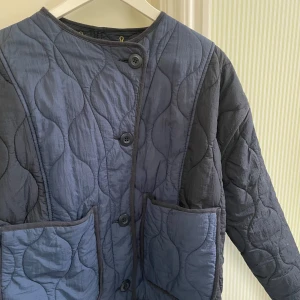 Blå quiltad jacka från Zara - Säljer en blå quiltad jacka från Zara med stora fickor framtill och knäppning med knappar. Jackan har en rund halsringning och långa ärmar. Perfekt för en stilren look. 