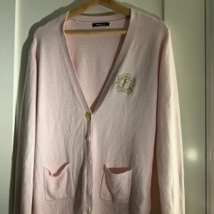 Rosa kofta från Gina Tricot - Säljer en söt rosa kofta från Gina Tricot med guldfärgade knappar och ett broderat emblem på bröstet. Koftan har långa ärmar och två fickor framtill. Perfekt för en stilren och bekväm look, men några knappar saknas 