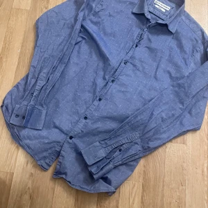 Blå skjorta från The Shirt Factory 42 - Snygg blå skjorta från The Shirt Factory 42 i slim fit. Skjortan är tillverkad i 100% bomull och har ett diskret mönster med små kryss. Perfekt för en stilren look med knappar framtill och långa ärmar.