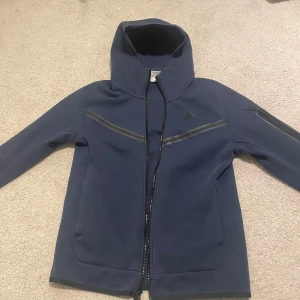 Mörkblå jacka från Nike - Säljer en mörkblå jacka från Nike med dragkedja och hög krage. Jackan har en stilren design med långa ärmar och praktiska fickor med dragkedjor. Perfekt för en sportig look eller vardagsbruk. Funkar för många storlekar