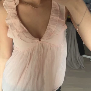 Ljusrosa blus med volangdetaljer - Säljer en söt ljusrosa blus med volangdetaljer vid halsringningen. Blusen har en djup v-ringning och är ärmlös, vilket gör den perfekt för varmare dagar. Använd max 2 ggr, Från Gina tricot! Skriv för frågor💕