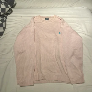 Ljusrosa stickad tröja från Ralph Lauren - Säljer en ljusrosa stickad tröja från Ralph Lauren med en liten blå logga på bröstet. Tröjan har långa ärmar och ribbade muddar vid ärmslut och nederkant. Perfekt för en stilren och bekväm look.