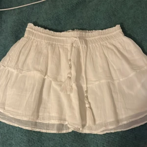 Vit kortkjol med volang - Säljer en söt vit kortkjol med volangdetaljer och knytband i midjan. Perfekt för varma sommardagar. Kjolen har en luftig och lätt design som ger en avslappnad känsla.