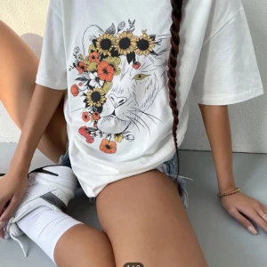 Vit t-shirt med lejonmotiv och blommor - Säljer en  vit t-shirt med ett stort lejonmotiv täckt av färgglada blommor. Den är perfekt för sommaren och har en avslappnad passform. Materialet är mjukt och bekvämt, perfekt för vardagsbruk. 