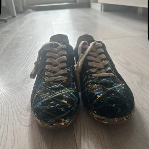 Färgglada sneakers från Maison Margiela - Säljer ett par unika sneakers från Maison Margiela i storlek 42. Skorna har en svart bas med ett coolt färgstänk-mönster i blått, gult och vitt. De har beige snörning och en brun sula. Perfekta för att ge din outfit en konstnärlig touch!