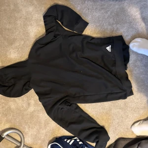 Svart hoodie från Adidas - Säljer en svart hoodie från Adidas i mycket bra skick. Den har en dragkedja framtill och Adidas-loggan tryckt på sidan. Perfekt för träning eller en chill dag. Den är långärmad och har en skön passform. Hör av dig om du är intresserad!