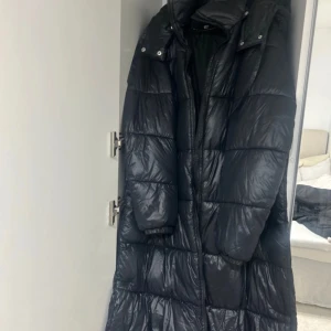 Svart lång pufferjacka från 157 - Säljer en svart lång pufferjacka från 157 i mycket bra skick. Jackan är perfekt för kalla vinterdagar och har en skön, vadderad design med dragkedja och knappar. Den är långärmad och har en hög krage för extra värme. Perfekt för både vardag och vinteräventyr!