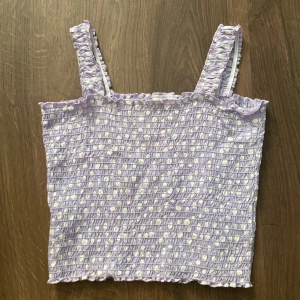 Lila topp med prickar/ blommor  från Kappahl - Söt lila topp från Kappahl med små vita blommor med en gul prick i. Den har en smockad design och breda axelband. Perfekt för sommardagar och kan enkelt matchas med jeans eller shorts. Toppen är i mycket bra skick och redo för nya äventyr!