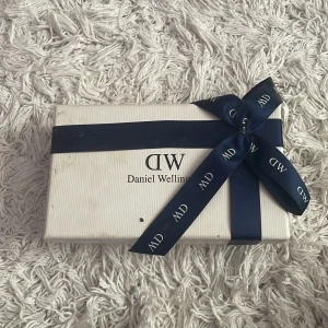 Daniel Wellington klocka med randigt armband - Säljer en stilren Daniel Wellington klocka med ett randigt armband i blått, vitt och rosa. Klockan har en klassisk vit urtavla och silverfärgad boett. Perfekt för både vardag och fest! Kommer i originalförpackning med blått band.