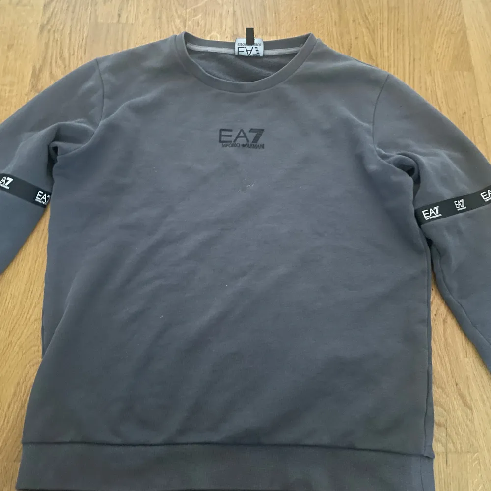 Säljer en grå sweatshirt från Emporio Armani EA7. Tröjan har en klassisk rund halsringning och långa ärmar med EA7-logga på ärmarna. Perfekt för en casual look eller träning. Den är i mycket bra skick. Fläcken i mitten är bara en lite som kan tvättas bor med vatten om jag ska fixa!. Tröjor & Koftor.