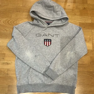 Grå hoodie från GANT - Säljer en snygg grå hoodie från GANT med deras klassiska logga och ett emblem på bröstet. Perfekt för både vardag och chill. Den har en skön passform och är i bra skick, dock sliten vid armbågarna. Perfekt för höst och vår!