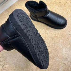 Svarta boots UGZ - Säljer ett par svarta boots i syntetmaterial med mjukt fuskpälsfoder. Perfekta för kalla dagar med sin varma insida. De har en robust sula för bra grepp och en stilren design som passar till det mesta. Märket UGZ JXH9DS syns på baksidan. Perfekta för vintern!