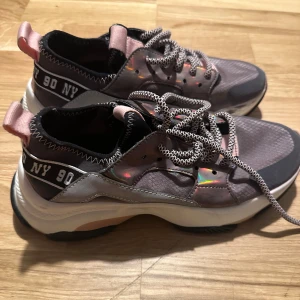 Steve Madden sneakers med rosa detaljer - Säljer ett par Steve Madden sneakers i grått med coola rosa detaljer och snörning. De har en chunky sula och texten '90 NY' på sidan. Perfekta för en trendig look!