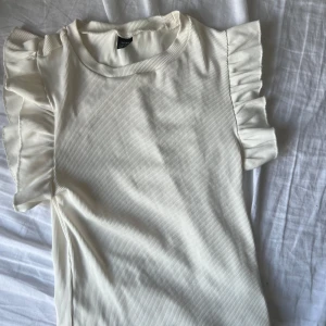 Vit ribbad topp från Niay - Säljer en superfin vit ribbad topp från Niay i storlek XS. Den har korta ärmar med volangdetaljer som ger en söt touch. Perfekt för både vardag och festliga tillfällen. Toppen är i mycket bra skick och redo för en ny garderob!