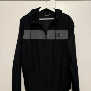 Svart vindjacka från Under Armour - Säljer en svart vindjacka från Under Armour i storlek M. Jackan har en grå detalj över bröstet och en dragkedja framtill. Perfekt för träning eller som en lätt jacka för höstvädret. Den har en huva och är i mycket bra skick.