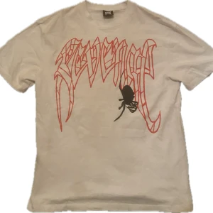 Revenge Tshirt - Säljer en beige t-shirt från Revenge med ett coolt tryck. Framsidan har en röd text och en svart spindel, medan baksidan har en röd dödskalle med ett 'R' i mitten. Perfekt för en avslappnad stil eller fest. Skön och bekväm att bära! TRÖJAN ÄR VIT IRL DÅLIGT LJUS