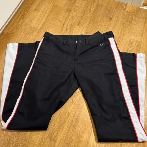 Svarta vintage byxor från Nike - Säljer ett par svarta vintage byxor från Nike i storlek 36/S . Byxorna har en vit och röd rand längs sidorna och är i mycket bra skick.