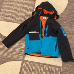 Blå och svart jacka från McKinley - Säljer en cool blå och svart jacka från McKinley i storlek 150. Den har en orange insida och är perfekt för utomhusaktiviteter. Jackan har dragkedja och flera praktiska fickor. Perfekt för höst och vinter!