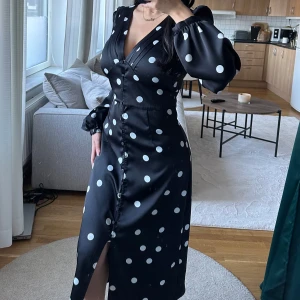 Svart prickig klänning - Snygg svart klänning med vita prickar och v-ringad design. Den har långa ärmar och en knappdetalj framtill som ger en elegant touch. Perfekt för både fest och vardag!