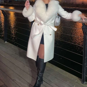 Vit kappa med pälsdetaljer - VALERIE kappa från femenina! Säljer en elegant vit kappa med fluffiga pälsdetaljer på krage och ärmslut. Kappan har ett knytbälte i midjan och är perfekt för kyliga kvällar. Den är långärmad och har en stilren design som passar både till vardags och fest.