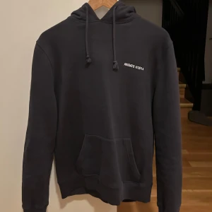 Svart hoodie från Axel Arigato - Säljer en stilren grå hoodie från Axel Arigato med tryck 'ARIGATO ©2014' på bröstet och ryggen. Den har en klassisk känguruficka och justerbar huva med dragsko. Perfekt för en avslappnad look eller kyliga dagar. 🖤