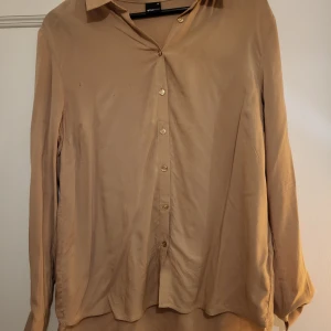 Beige blus från Gina Tricot - Säljer en stilren beige blus från Gina Tricot i storlek 38. Blusen har långa ärmar och knappar framtill. Perfekt för både vardag och fest! Den är i bra skick med några små märken, har en provat fläckborttagning på fläckarna. 
