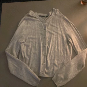 Grå kofta med knappar - Mysig grå kofta i mjukt material med knappar framtill. Perfekt för kyliga dagar och att ha över en topp. Den har långa ärmar och en klassisk passform. Ett måste i garderoben för höst och vinter!