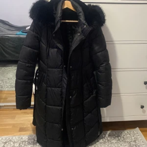 Svart dunjacka med pälsdetaljer - Säljer en svart lång dunjacka med mysig päls på huvan. Perfekt för kalla vinterdagar! Jackan har en snygg quiltad design och ett bälte i midjan för en fin passform. Den är långärmad och har både dragkedja och knappar framtill. Supervarm och stilren!