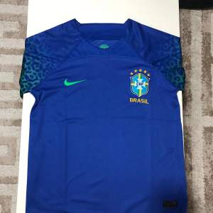 Säljer en blå Brasilien fotbollströja från Nike i Dri-FIT material. Tröjan har korta ärmar med ett coolt mönster i grönt och blått. Brasilien-emblemet och Nike-loggan är tryckta på framsidan. Perfekt för fotbollsfans som vill visa sitt stöd för Brasilien! 🌟perfekt skick nästan helt ny.