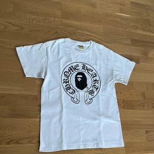 Bape x Chrome Hearts T-shirt  - Säljer en cool vit t-shirt från A Bathing Ape med tryck av Chrome Hearts och Baby Milo. T-shirten har korta ärmar och är i bomull, perfekt för en avslappnad stil. Den är i mycket bra skick och passar perfekt till vardags 