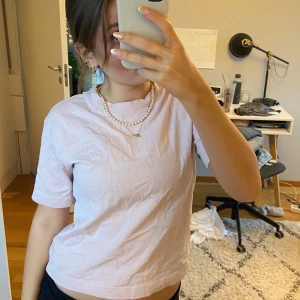 Ljusrosa t-shirt - Säljer en ljusrosa t-shirt i bomull. Den är kortärmad och har en croppad passform, perfekt för sommardagar. Enkel och stilren design som passar till det mesta. Perfekt för en avslappnad look!