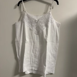 Vit linne med spetsdetaljer - Säljer ett sött vitt linne med justerbara axelband och vacker spetsdetalj framtill. Perfekt för sommardagar eller som ett lager under en tröja. Linnet är i bra skick och har en liten rosett i mitten av spetsen för en extra touch.